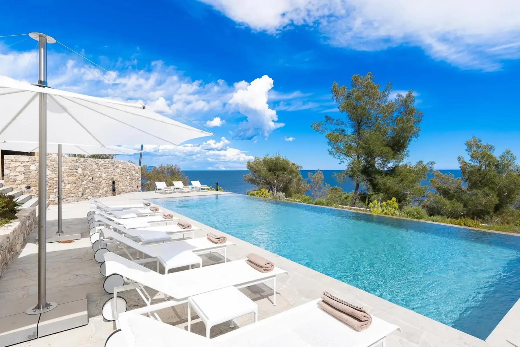Piscina infinita al aire libre con tumbonas blancas y sombrillas, con vistas al océano, rodeada de árboles y vegetación bajo un cielo azul con nubes dispersas.