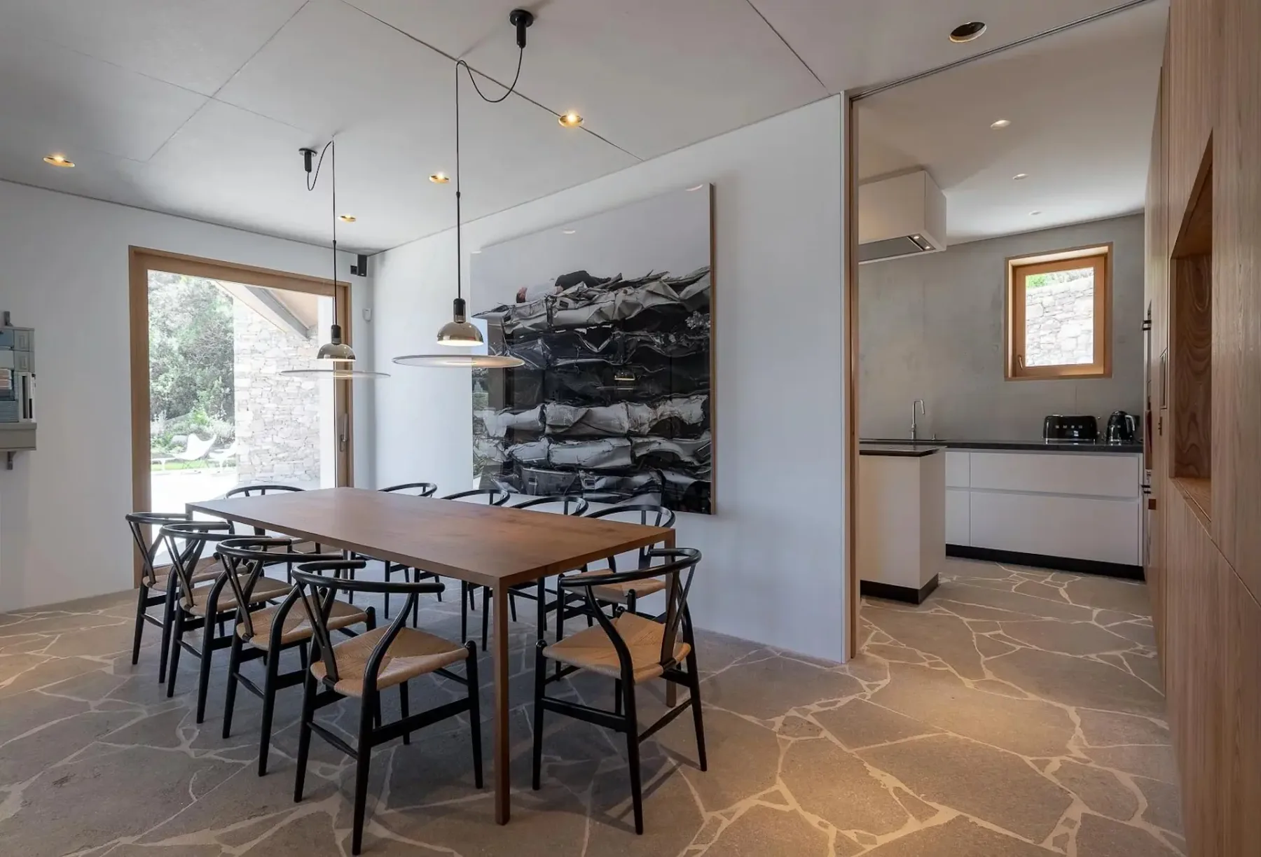 Un comedor moderno con una mesa de madera y sillas negras junto a una cocina contemporánea. El suelo es de baldosas de piedra y hay un cuadro grande colgado en la pared.