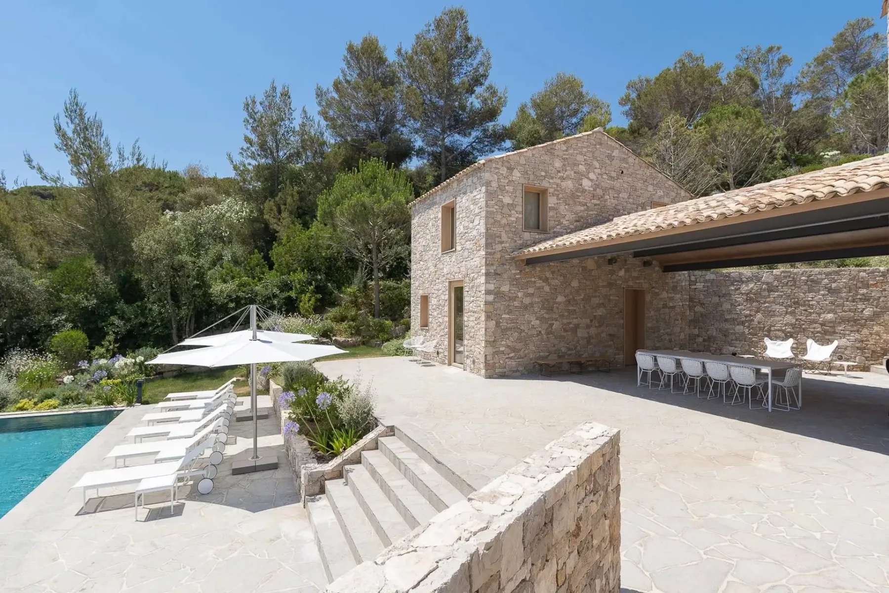 Una casa de piedra con un patio amplio, tumbonas blancas y sombrillas junto a una piscina, con árboles al fondo. A la derecha hay una zona de comedor exterior cubierta.