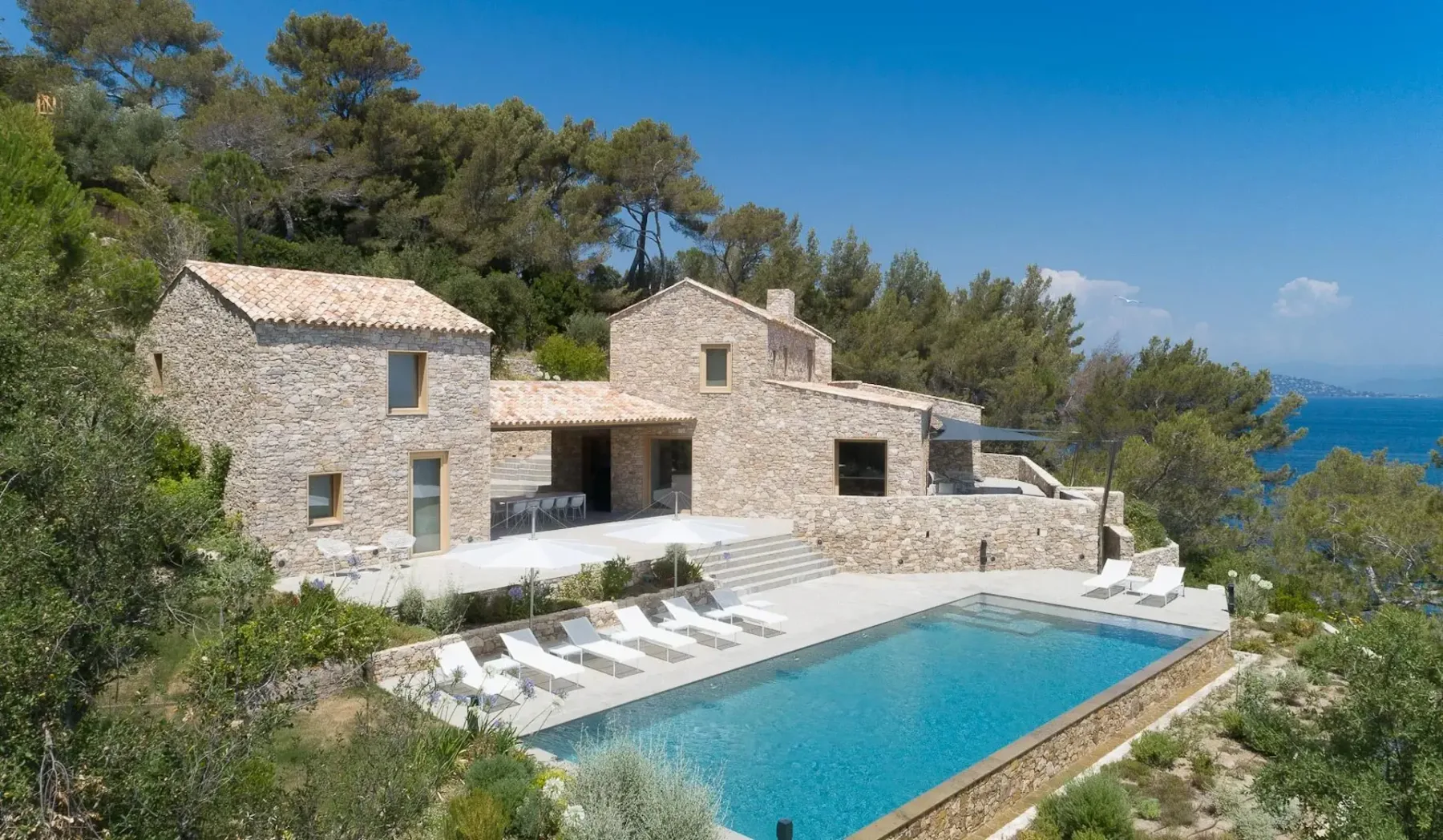 Una casa de piedra con tejado de tejas cuenta con una piscina al aire libre, tumbonas y una terraza, rodeada de árboles y con vistas a una masa de agua.