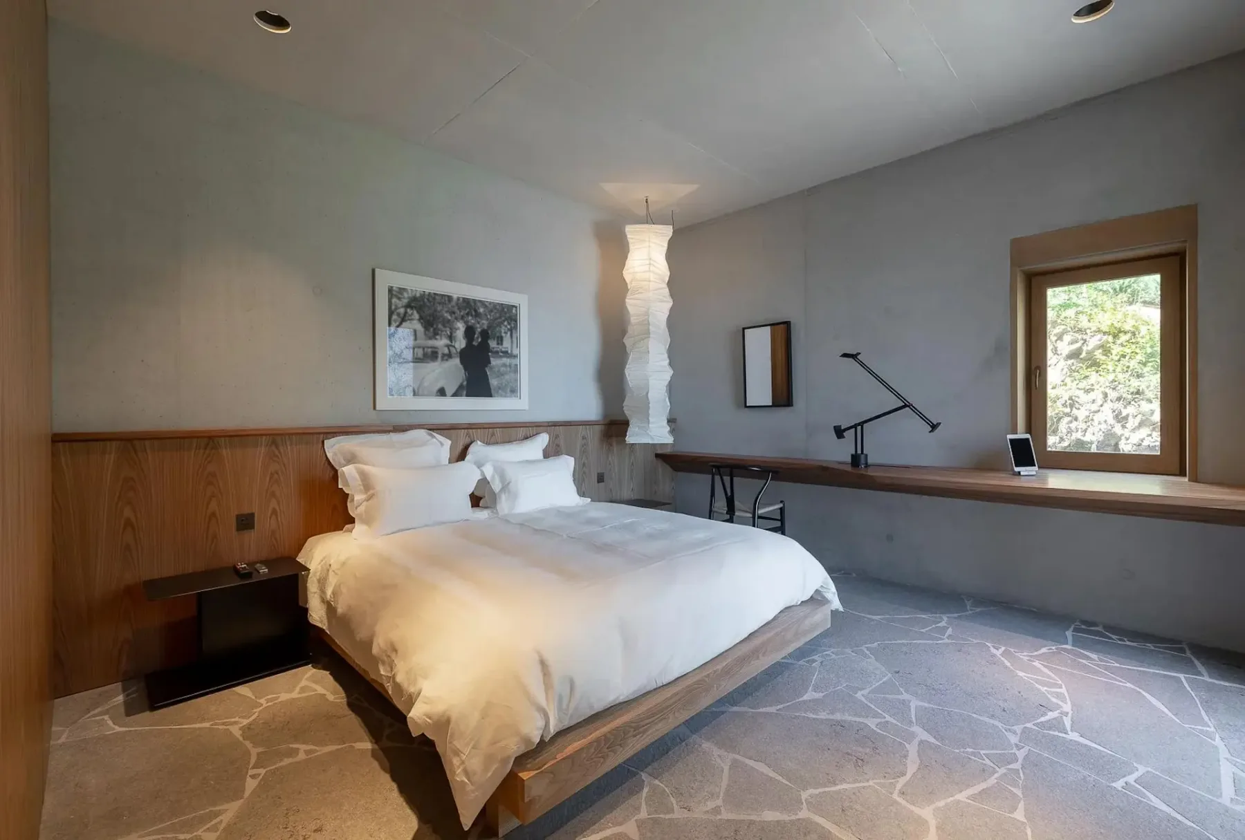 Dormitorio minimalista con una cama grande, ropa de cama blanca, un cabecero de madera, suelo de piedra, un escritorio fijado a la pared con una lámpara moderna y una tableta, una ventana y un singular farol colgante.