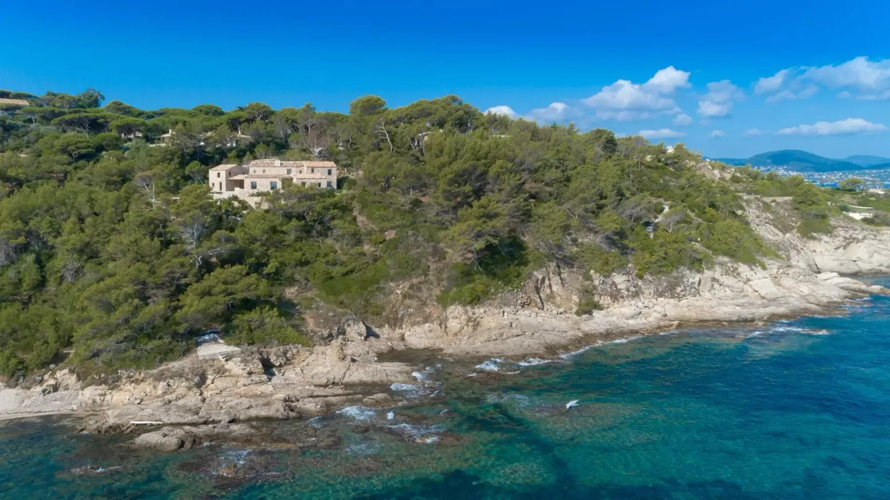 Vista aérea de una zona costera con una gran casa rodeada de vegetación densa sobre un acantilado rocoso con vistas al mar azul de aguas cristalinas.