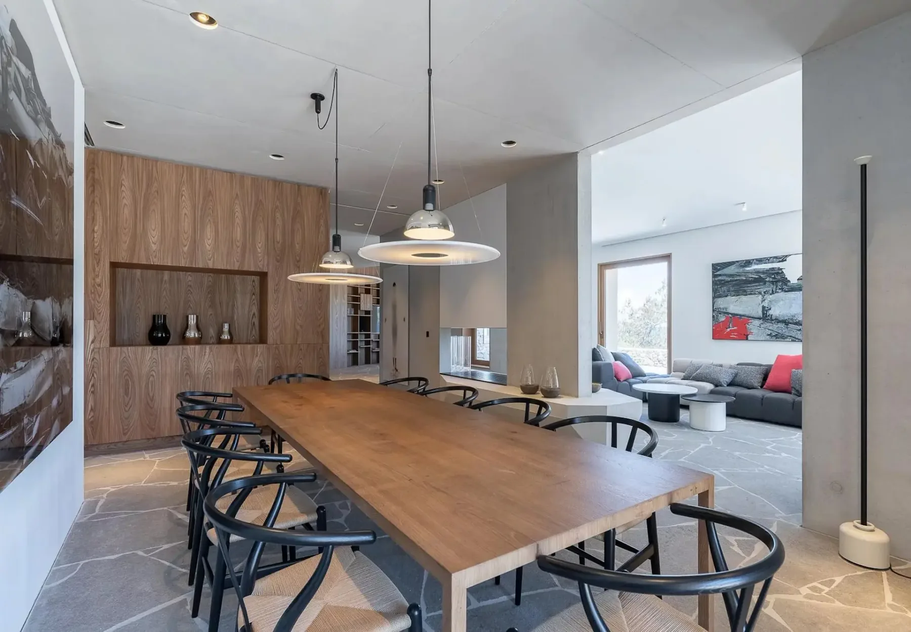 Un comedor moderno con una mesa larga de madera, sillas negras y lámparas colgantes, junto a una zona de estar con un sofá gris y arte abstracto en la pared. El suelo es de baldosas de piedra.