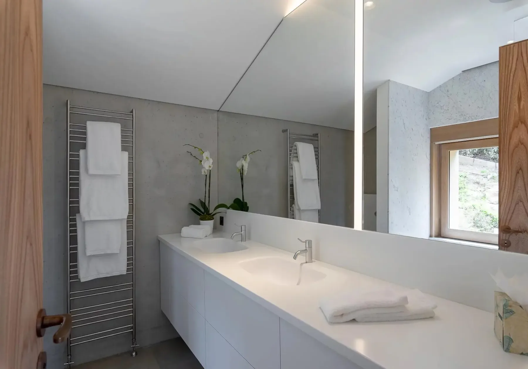 Baño moderno con un mueble largo y blanco con dos lavabos, un espejo grande, un toallero de pared, orquídeas en macetas y una ventana que deja entrar luz natural.