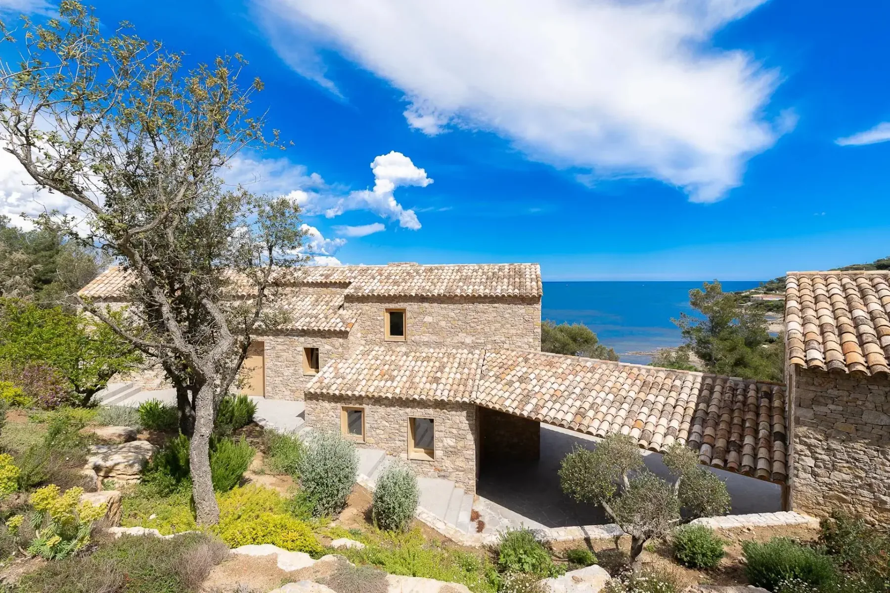 Una casa con tejado de tejas está entre árboles y arbustos, con el mar visible detrás.