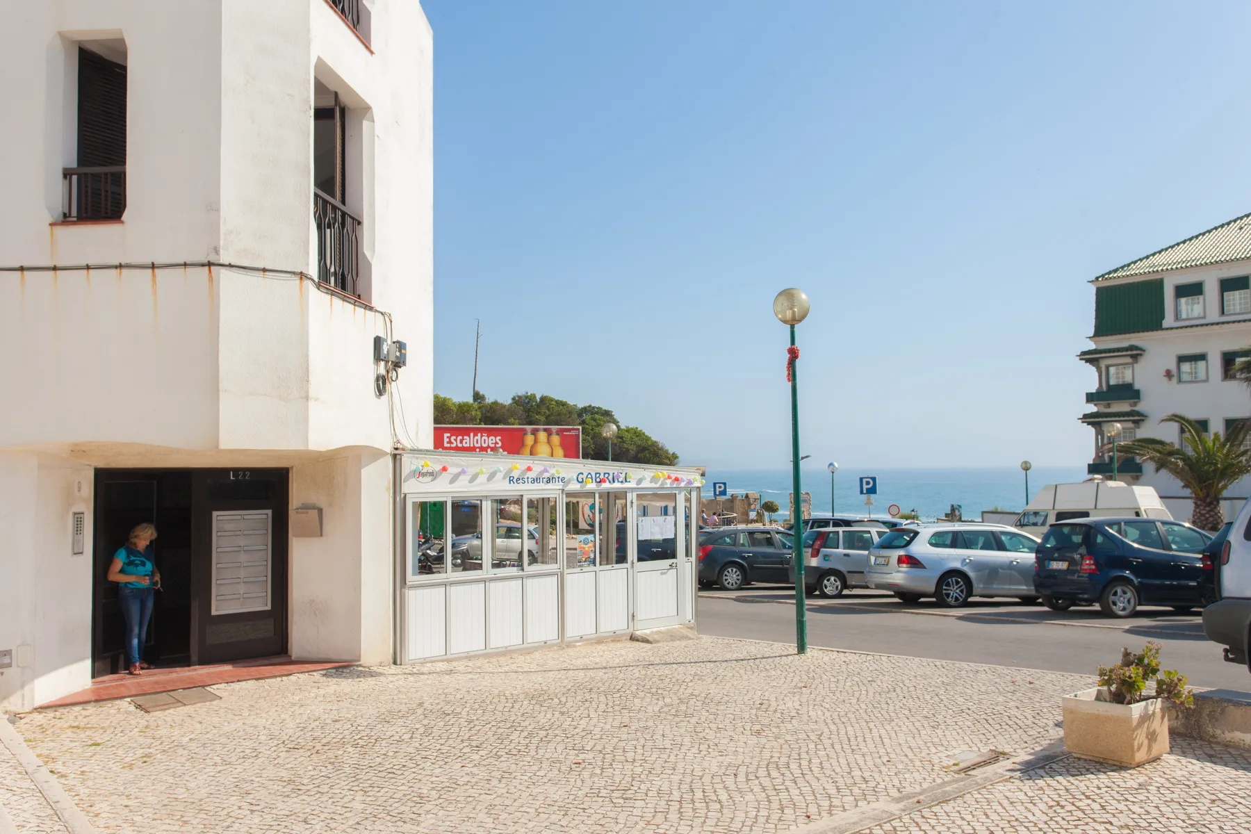 Una calle empedrada junto a edificios blancos, con coches aparcados y el océano visible a lo lejos.