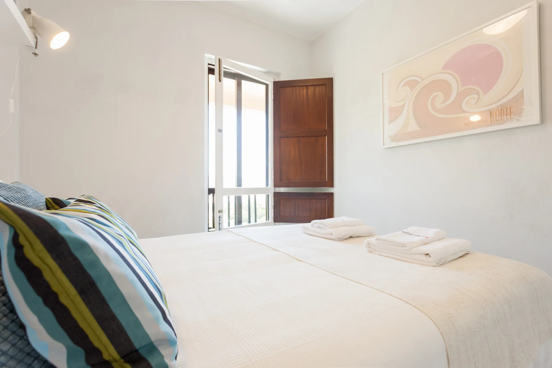 Un dormitorio con ropa de cama blanca, toallas dobladas y un cojín de rayas de colores.