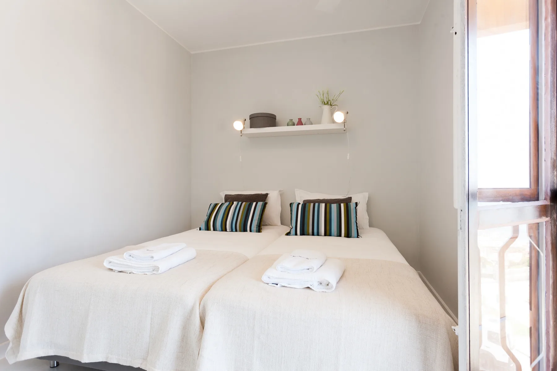 Un dormitorio luminoso con dos camas y almohadas a rayas bajo una pequeña repisa.