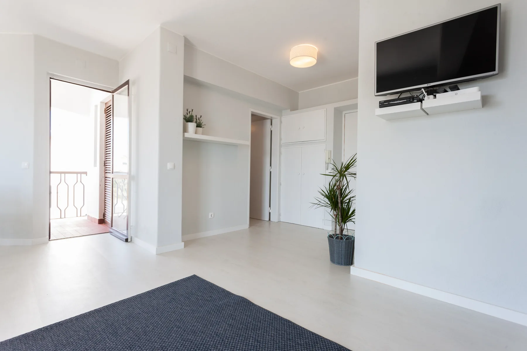Una sala luminosa con un televisor montado en la pared y una puerta abierta al balcón.