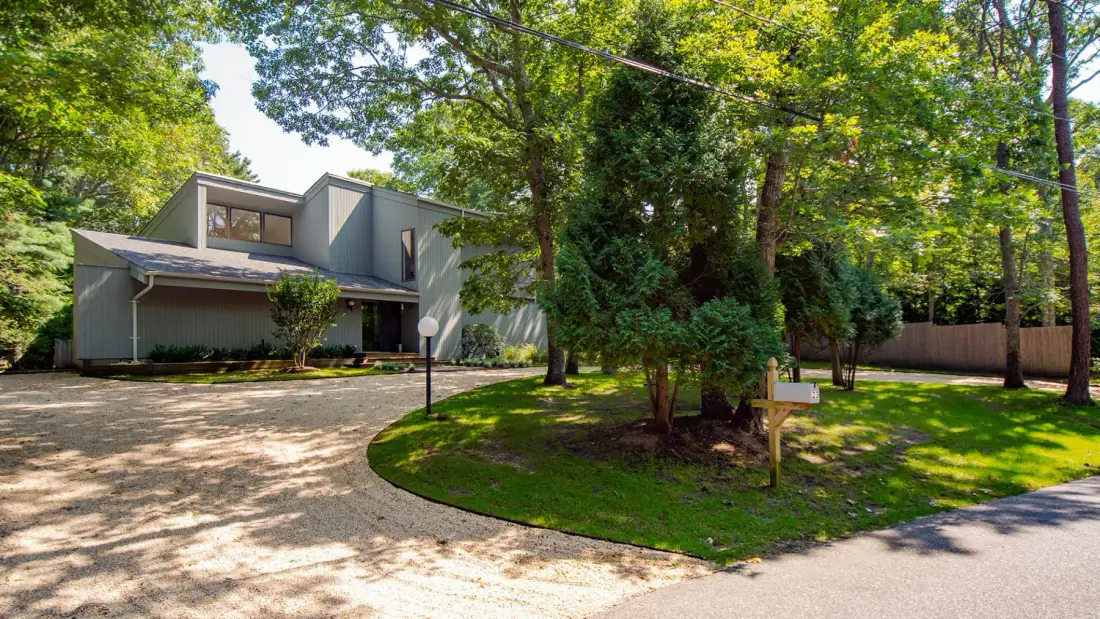 A modern house sits beside a curved driveway under leafy trees.