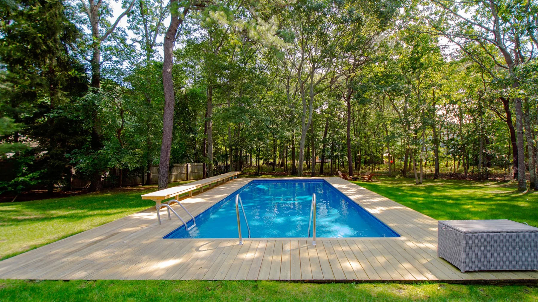 An outdoor swimming pool sits on a wooden deck amid leafy trees.