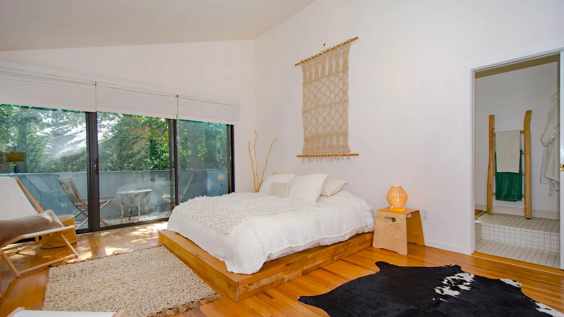 A bright bedroom with a bed, bedside table, and large windows overlooking greenery.
