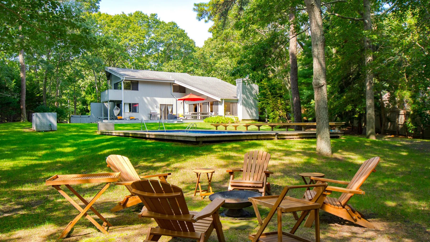 A modern house sits beyond a wooden deck, with outdoor seating on the lawn.