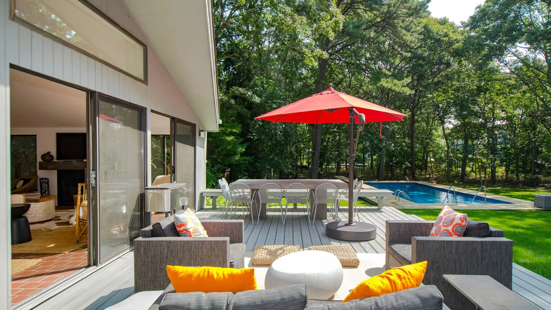 A patio with outdoor seating and a red umbrella overlooks a swimming pool.