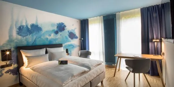 Habitación de hotel con una cama doble, un escritorio y ventanas grandes.