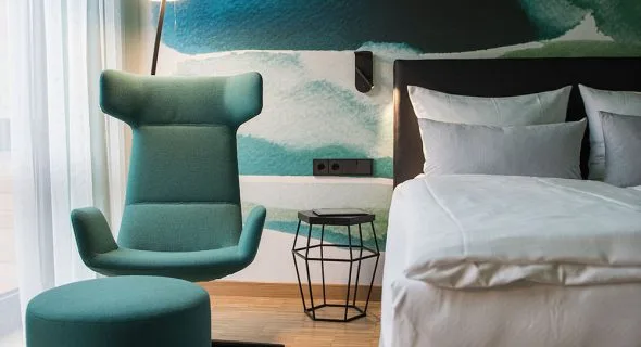 Habitación de hotel con una cama, una silla verde, suelo de madera y una ventana grande.