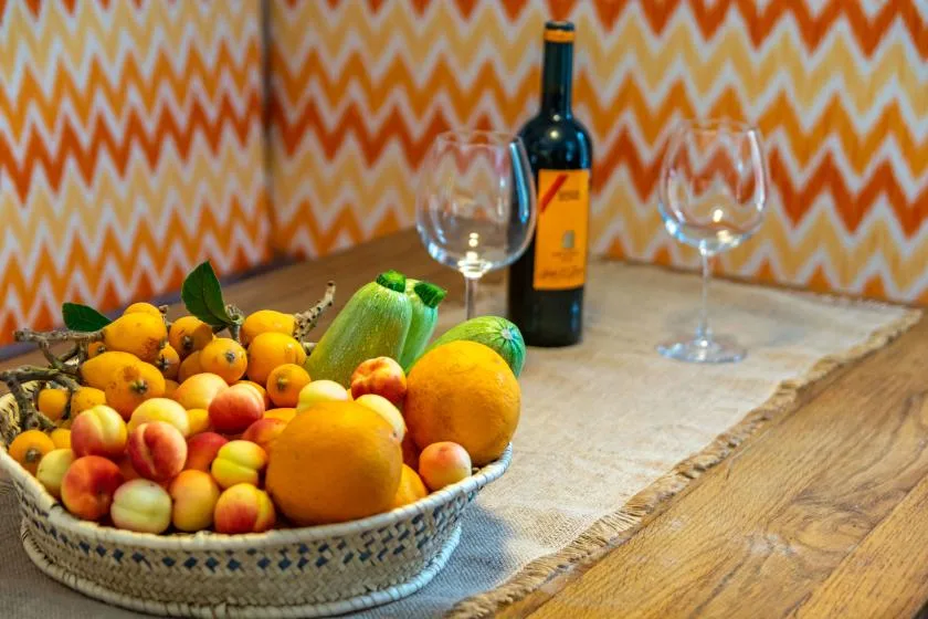A fruit bowl sits on a wooden kitchen counter beside a bottle and glasses.