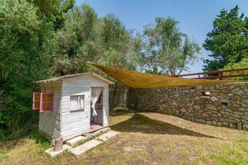 A small wooden garden shed sits beside a stone wall beneath a shade canopy.