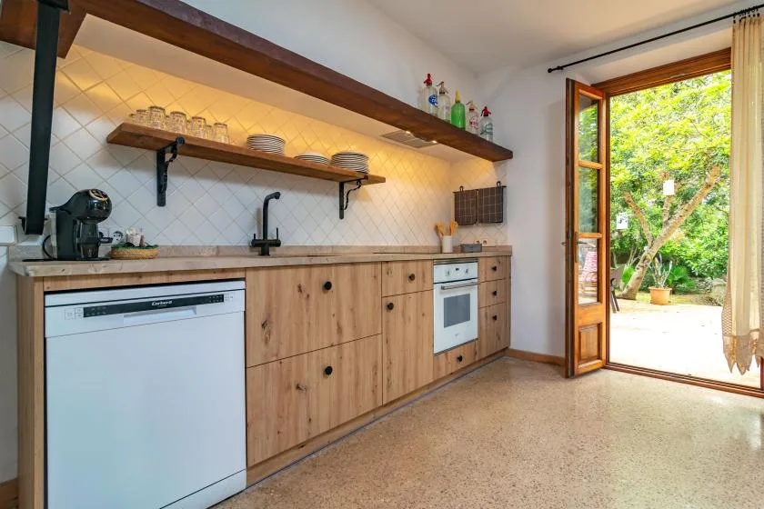 A bright kitchen with wooden cabinets, a counter, and an open door to greenery.