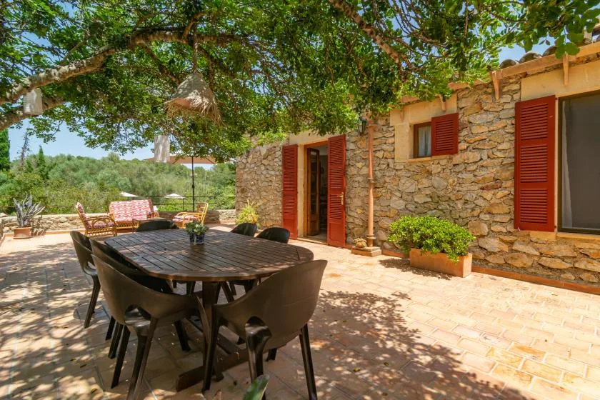 A shaded outdoor dining area beside a stone building, with a long table and seating.