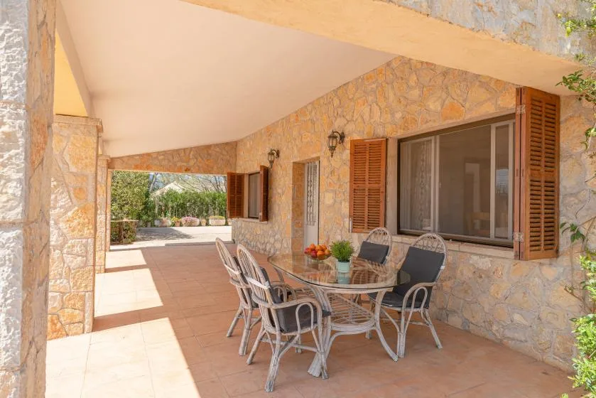Un patio cubierto con una mesa de comedor de vidrio junto a paredes de piedra y contraventanas abiertas.