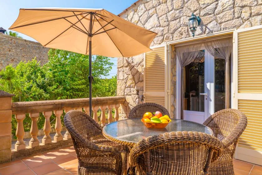 Una terraza de piedra tiene asientos de mimbre alrededor de una mesa de vidrio bajo una sombrilla grande.
