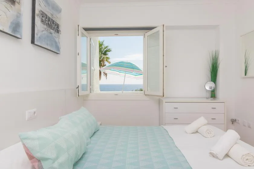 A bright bedroom with a bed beside an open window overlooking the sea.