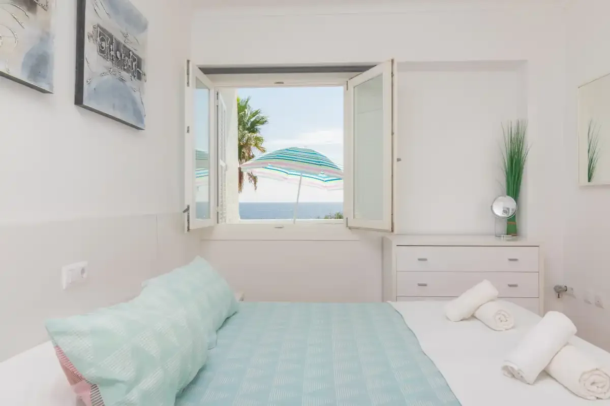 A bright bedroom with an open window overlooking the sea.