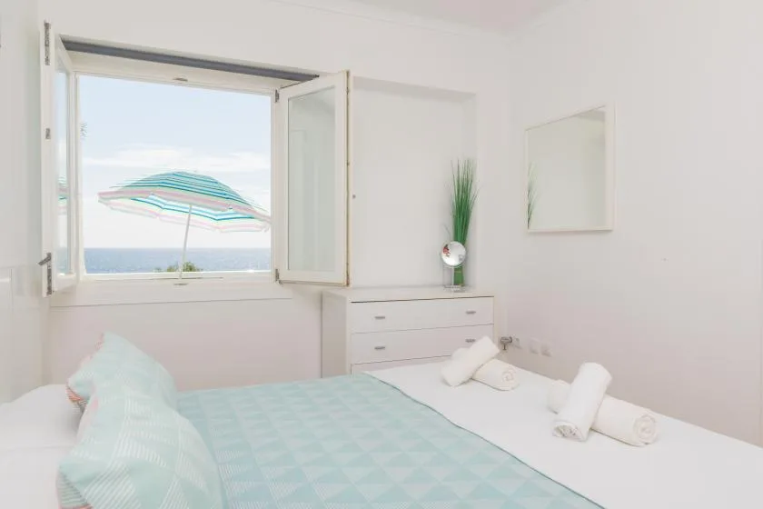 A bright bedroom with a bed beneath an open window overlooking the sea.