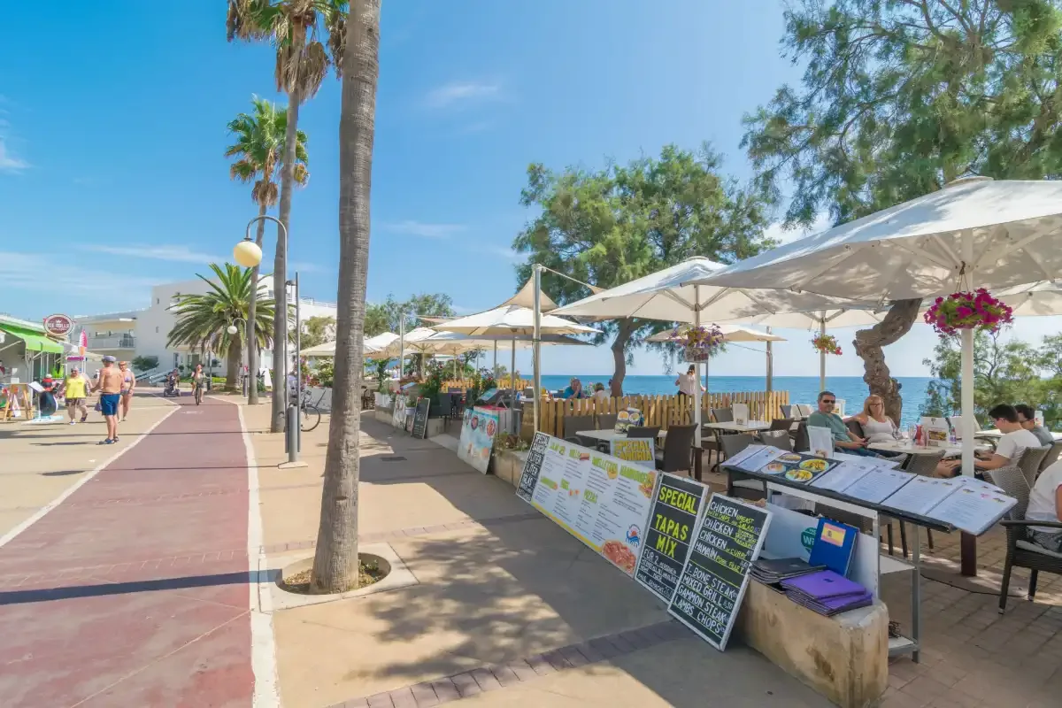 A seaside promenade with outdoor café tables beneath large umbrellas.