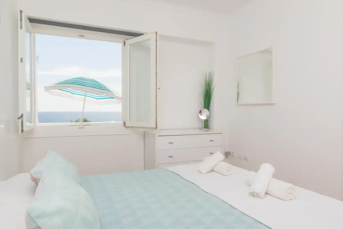 A bedroom with a bed beside an open window overlooking the sea.