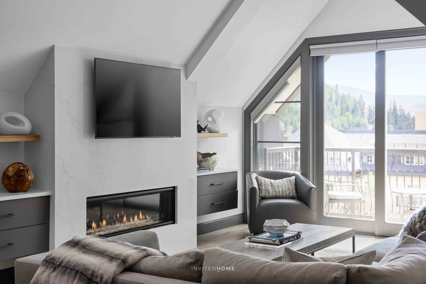 Una sala luminosa tiene una chimenea, un televisor en la pared y ventanas grandes.