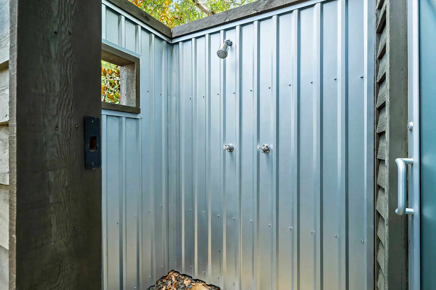 Ducha exterior con paredes de metal corrugado, un cabezal de ducha y dos mandos. Vista parcial de los árboles a través de una pequeña ventana rectangular en la parte superior.