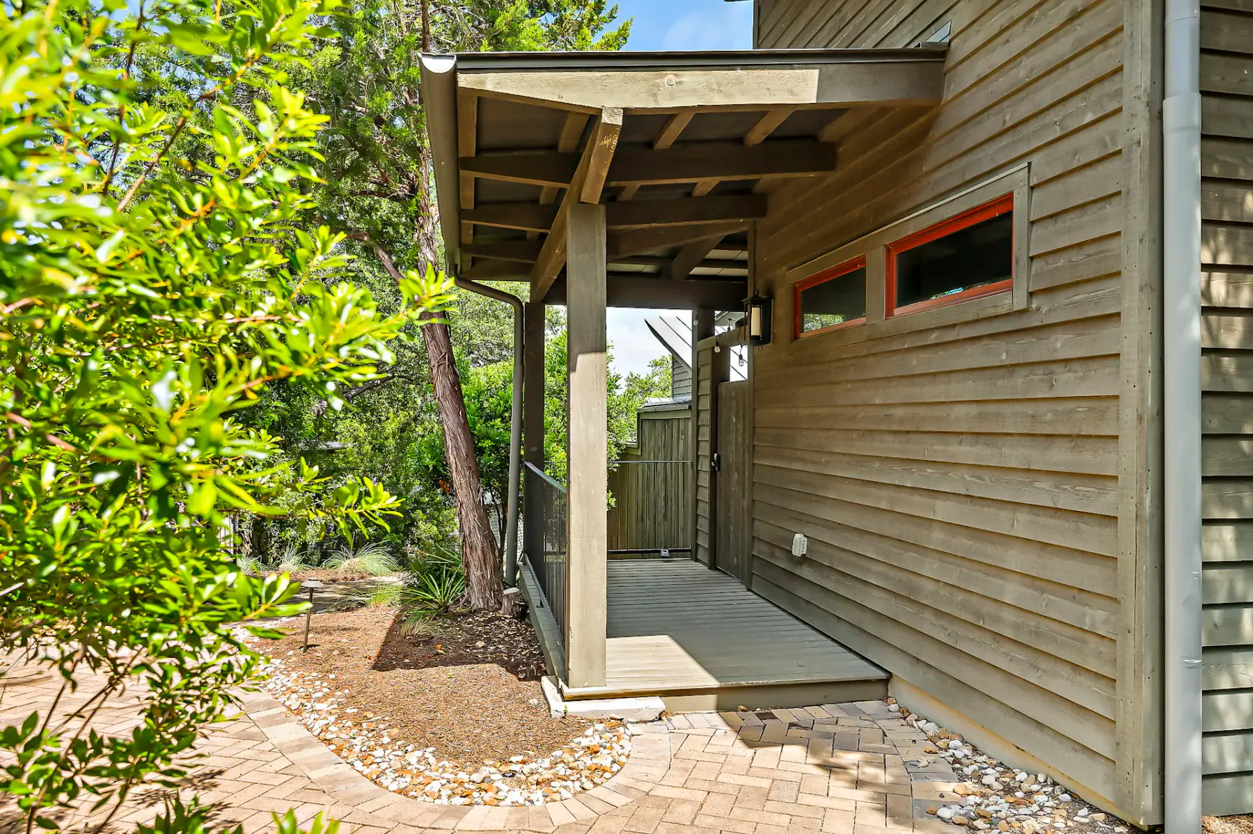Exterior de una casa de madera con un porche estrecho, rodeada de vegetación exuberante y un sendero de ladrillos bordeado de pequeñas piedras blancas.