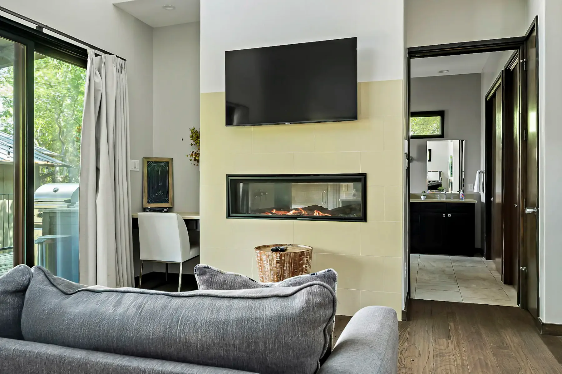 Sala de estar moderna con un sofá gris, un televisor montado en la pared sobre una chimenea y una zona de trabajo con una silla. Una puerta da paso a un baño luminoso.