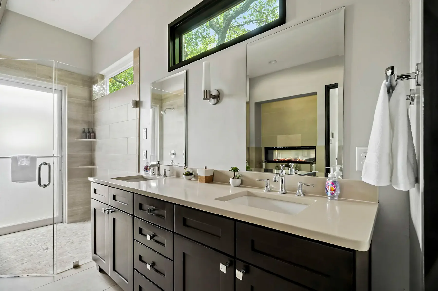 Baño moderno con dos lavabos, armarios de madera oscura, una ducha con cerramiento de vidrio y un espejo grande. Entra luz natural por ventanas altas.