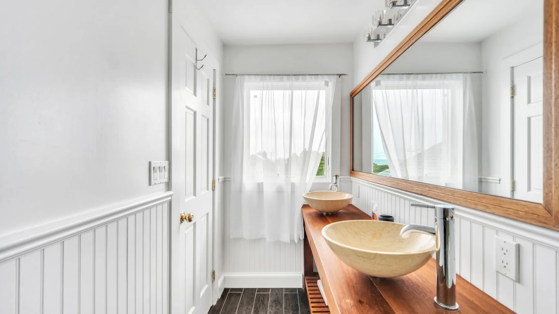 Un baño luminoso tiene un tocador largo de madera, un lavabo y cortinas translúcidas.
