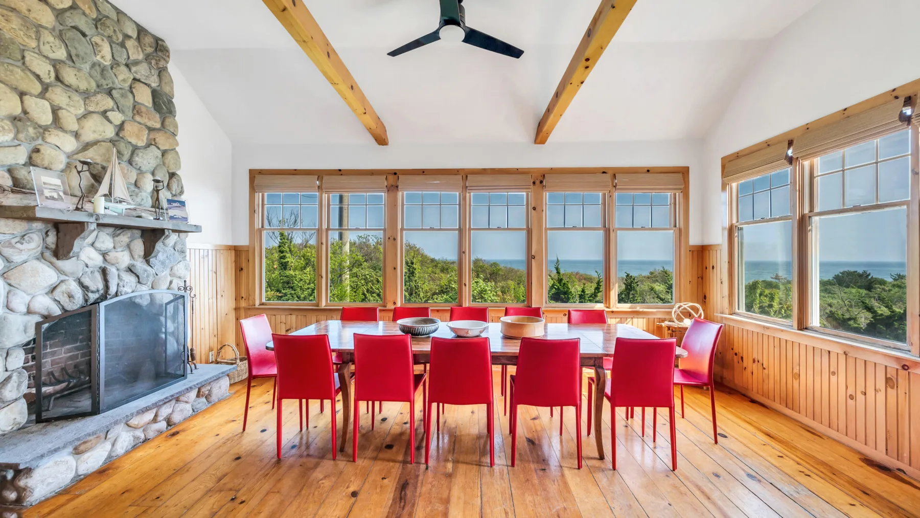 Comedor con sillas rojas alrededor de una mesa, junto a grandes ventanas y una chimenea de piedra.