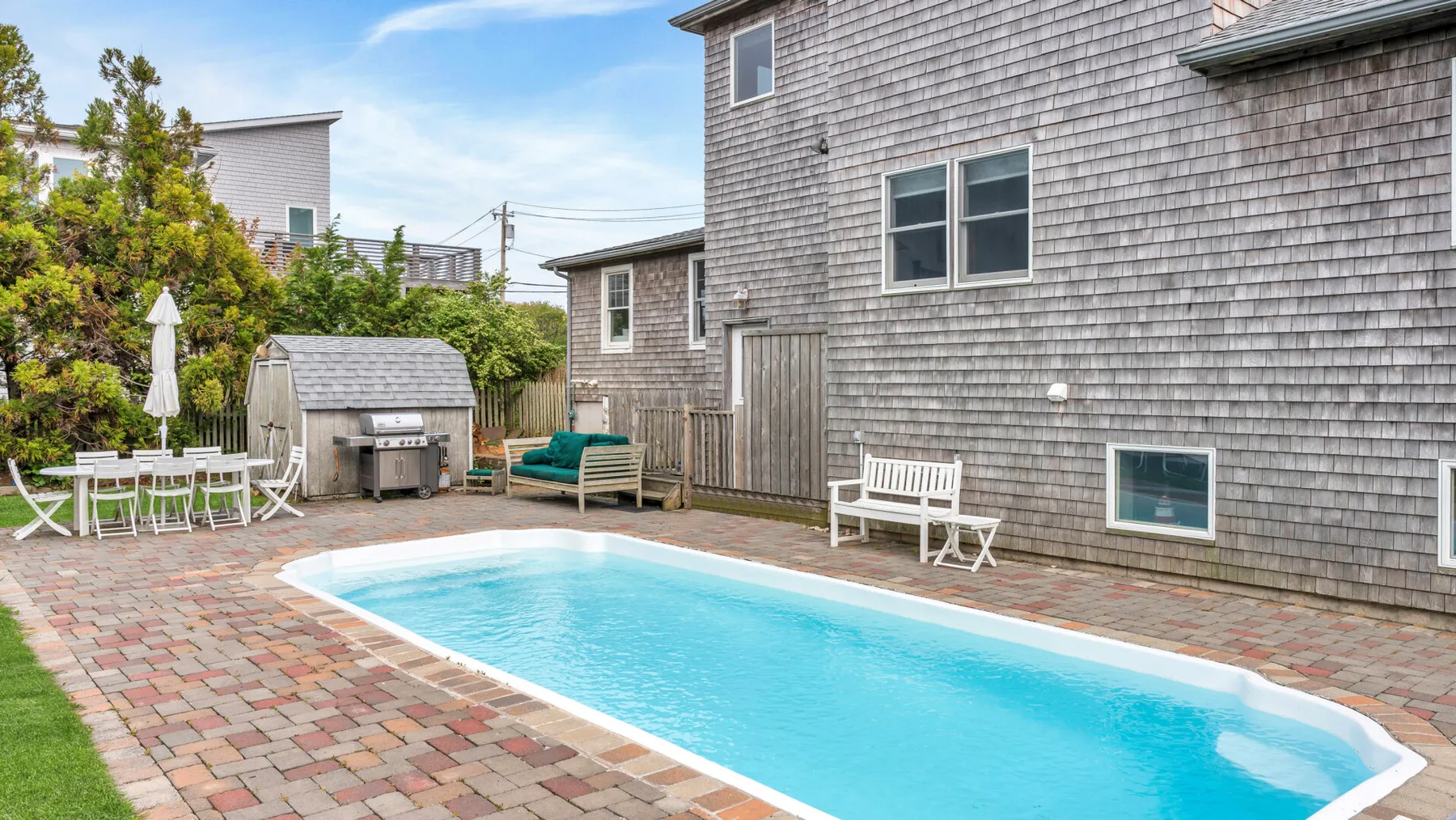 Una piscina está en un patio pavimentado junto a una casa revestida de tejas.