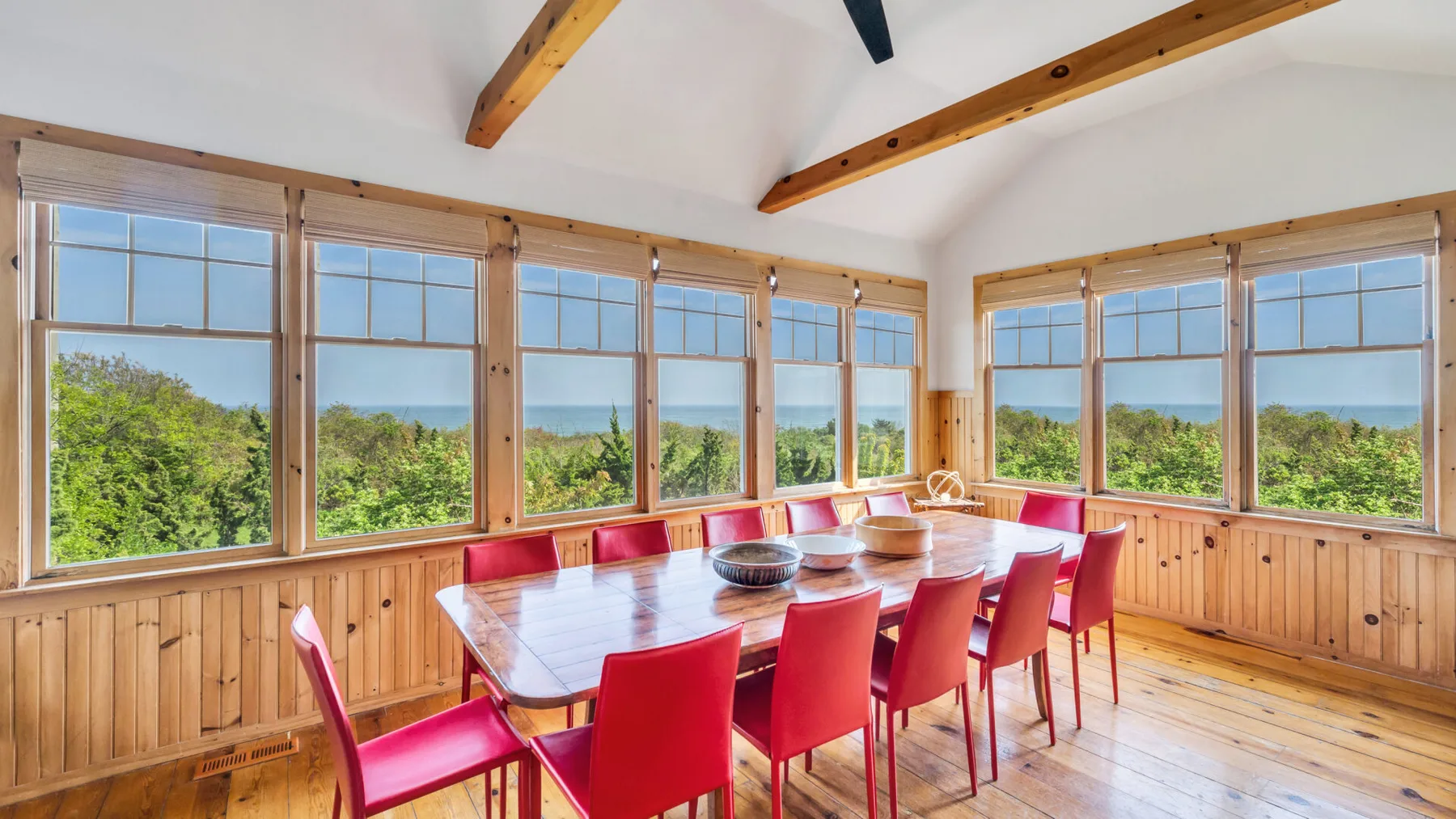 Un comedor con sillas rojas está junto a grandes ventanas con vistas a la vegetación y al agua.