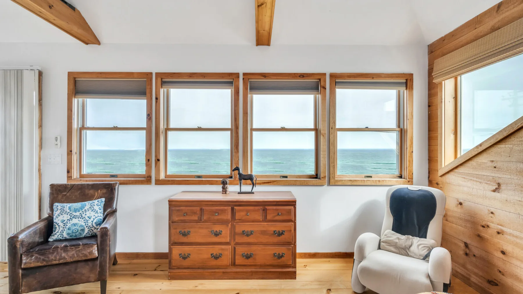 Un dormitorio luminoso con una cómoda de madera bajo cuatro ventanas y dos sillones.
