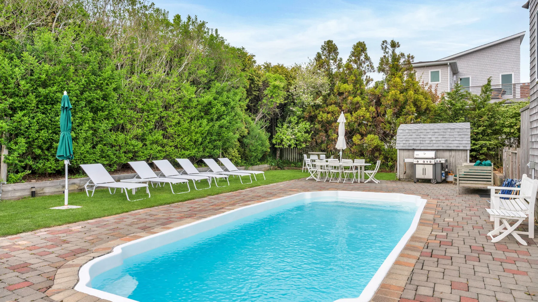 Una piscina al aire libre está junto a un patio pavimentado con asientos y vegetación.