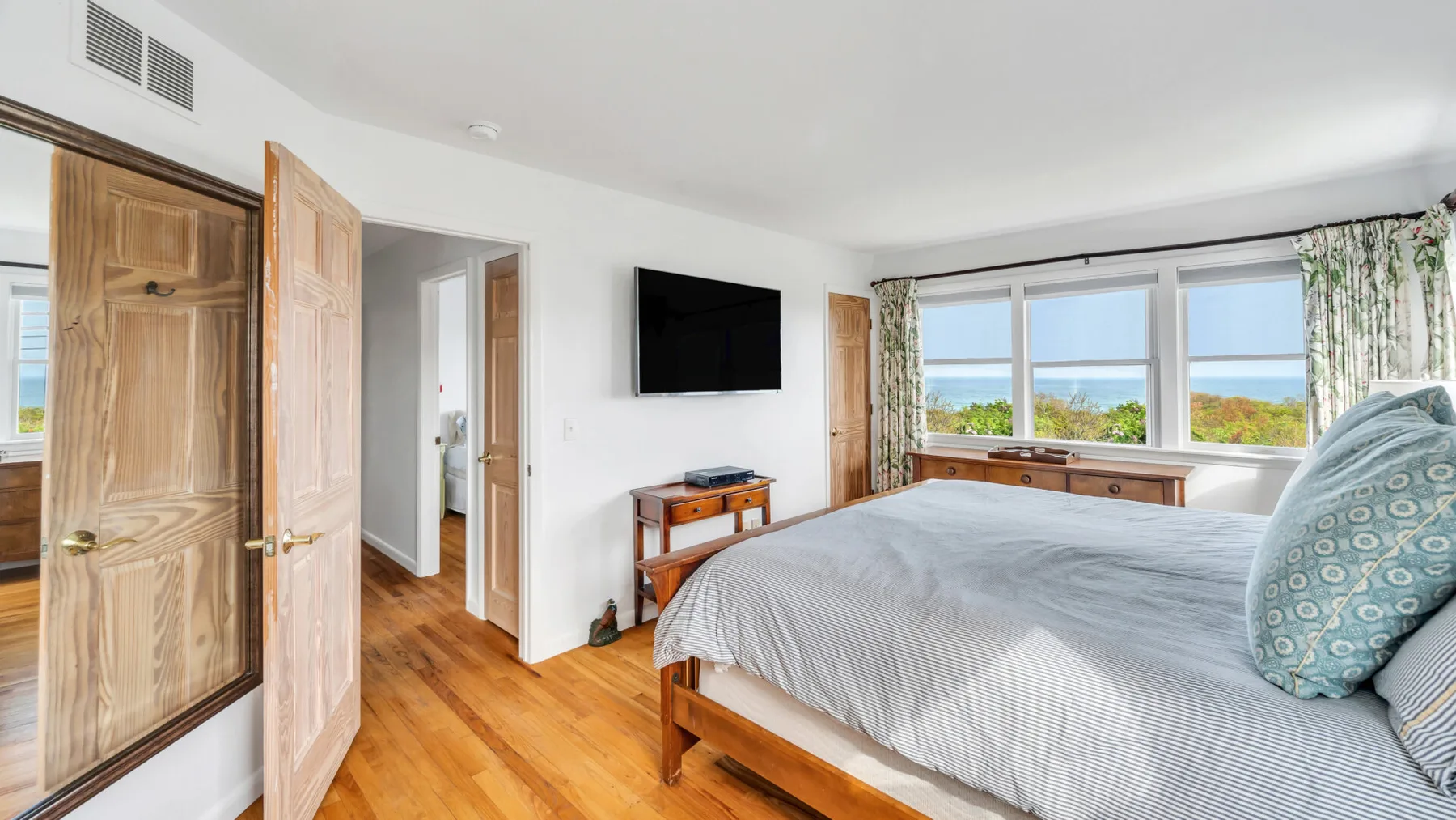 Un dormitorio con una cama grande, un televisor en la pared y ventanas con vistas a vegetación y agua.