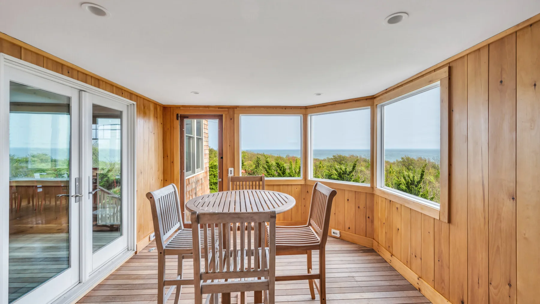 Un solárium revestido de madera tiene una mesa y grandes ventanas con vistas a la vegetación y al agua.