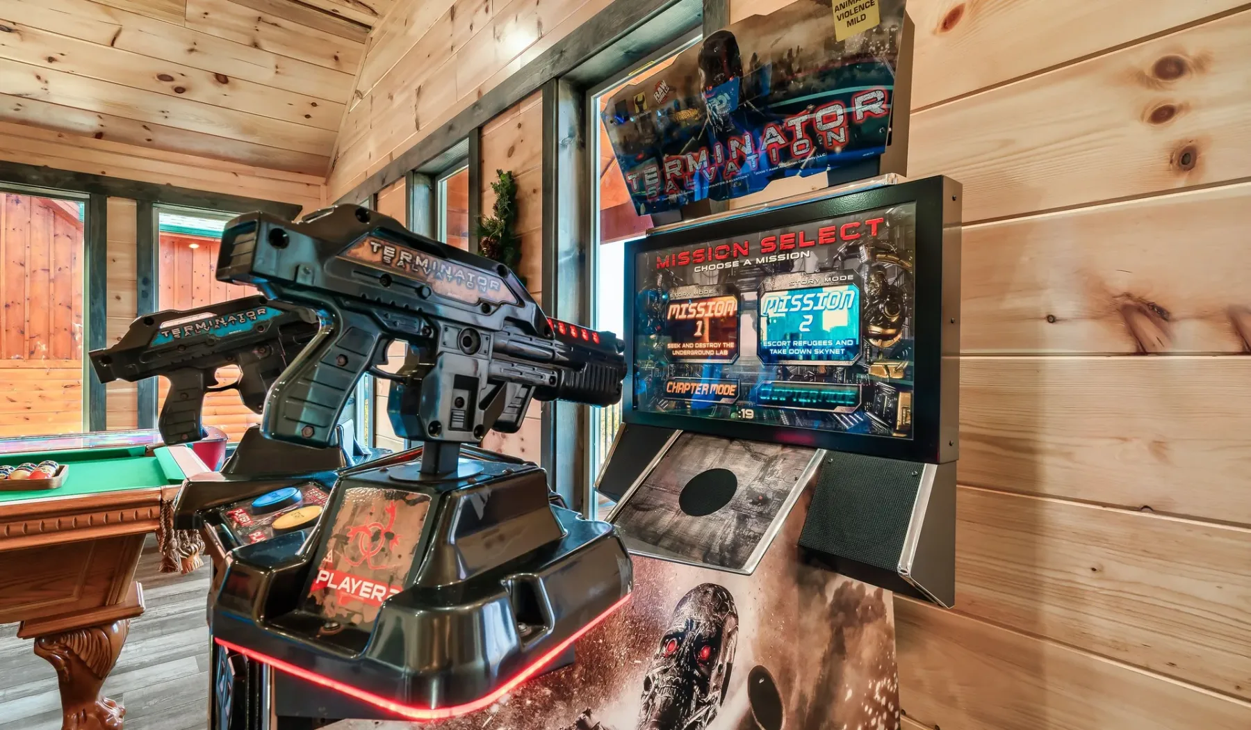 Una máquina recreativa con dos controles está junto a una mesa de billar en una sala revestida de madera.