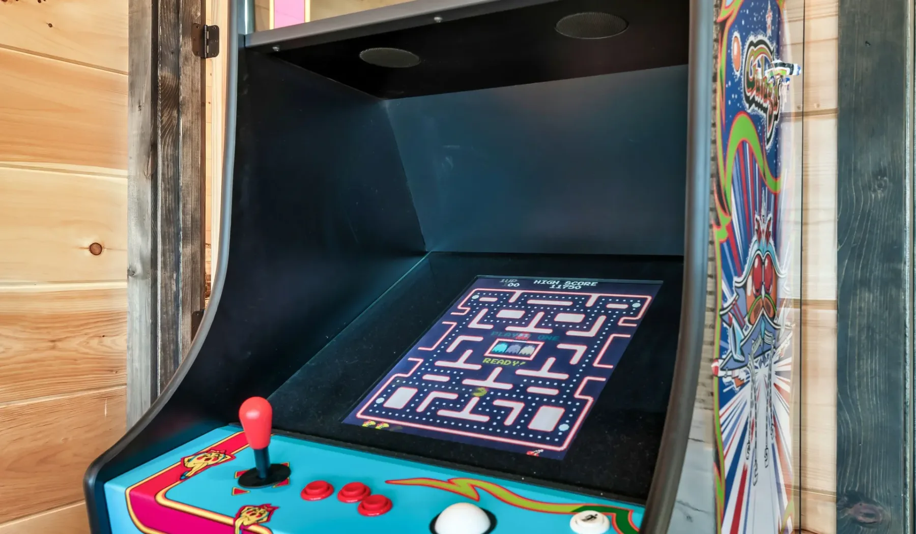 Una máquina recreativa con un panel de control colorido está junto a una pared de madera.