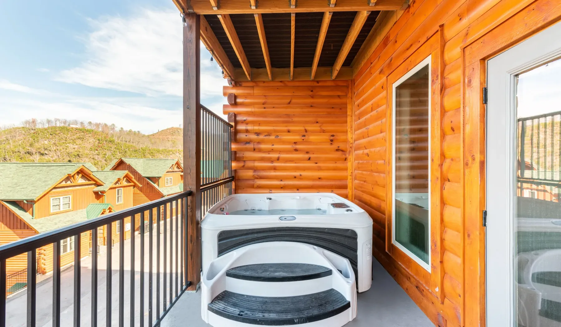 Un balcón cubierto de madera con una bañera de hidromasaje y vistas a cabañas cercanas.