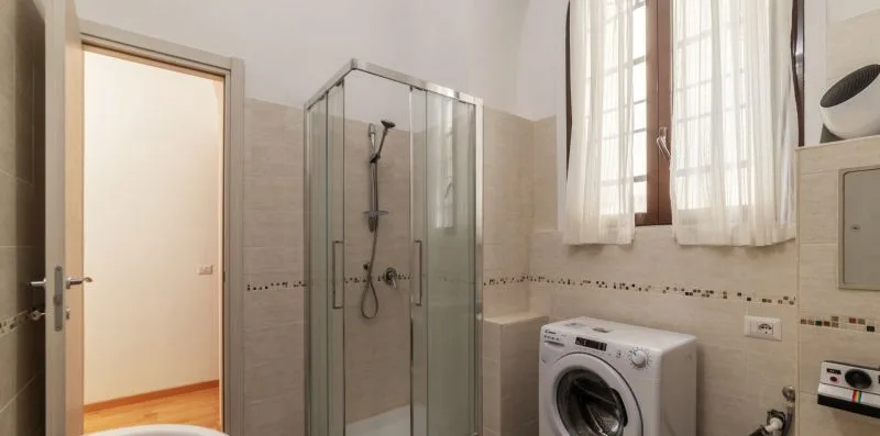 Un baño con ducha de vidrio, lavadora y una ventana luminosa.