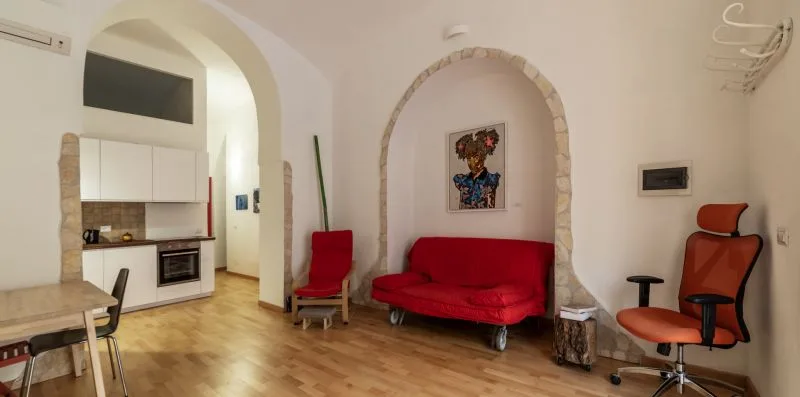 A living area with a red sofa, an orange office chair, and a compact kitchen.
