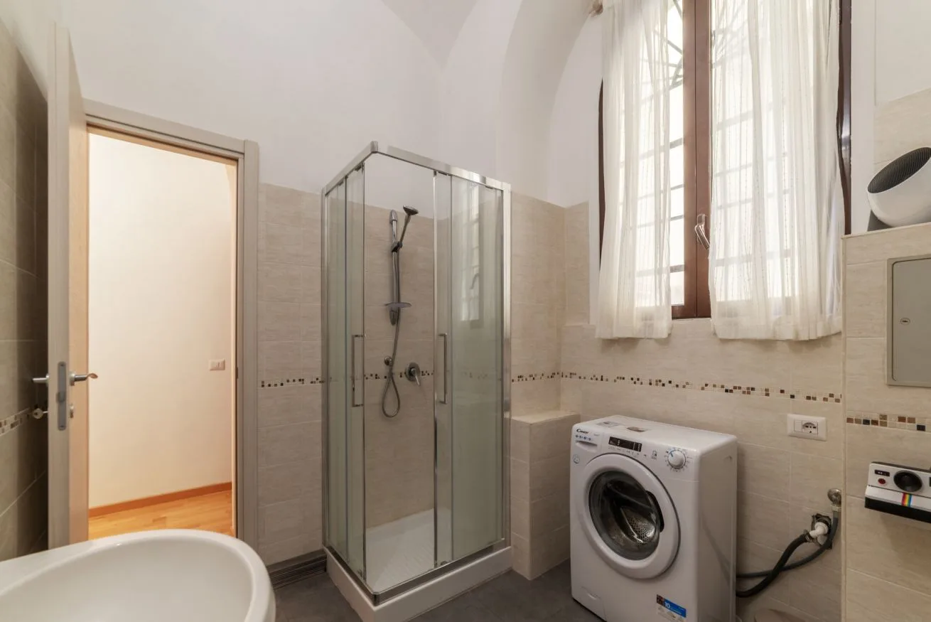 Un baño con ducha de vidrio, lavadora y una ventana alta.